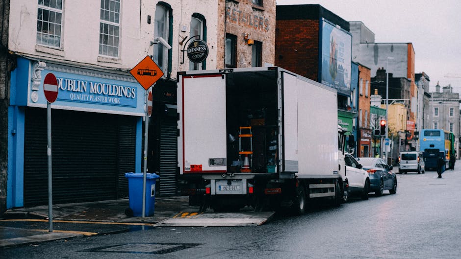 A white moving van is parked on a city street adjacent to a commercial building with a blue sign reading 'Dublin Mouldings' and a closed roller shutter. The rear doors of the van are open, revealing an interior storage area with visible equipment including orange securing straps and packing materials. The van is positioned on a yellow lined loading zone, with a blue recycling bin placed nearby on the pavement. Surrounding the van, there are several parked cars and a bus in the background, along with various storefronts and signage on multi-storey brick and concrete buildings. The scene appears to depict a home relocation process in an urban environment, with the loading process occurring outside a shop front under overcast weather conditions, illustrating typical furniture transport and packing activities associated with professional removals services such as those offered by Man with Van Shadwell.