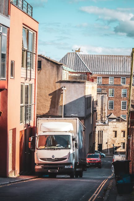 A white moving van parked on a narrow urban street, partially lodged between a pink multi-storey building on the left and a gray stone building in the background. The van is positioned with its front facing the viewer, and its rear cargo area is visible, indicating it is in the process of loading or unloading household furniture and boxes. Several cardboard boxes and wrapped furniture pieces can be seen inside the van's open rear doors, with some items also visible on the pavement nearby, possibly being prepared for transport or unpacked from packing materials like plastic wrap and blankets. The street is flanked by older, mixed-use buildings with brick and stucco facades, and the surrounding environment includes parked cars and a clear sky with some clouds. This scene illustrates home relocation activities involving furniture transport and packing during a professional removal process, as managed by Man with Van Shadwell.