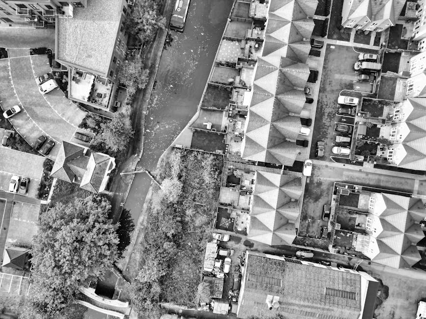 An aerial black-and-white photograph showing a residential street with row houses and terraced buildings, each with pitched roofs and small backyards. Moving vehicles and cars are parked along the street and on nearby driveways. A narrow, paved alley runs through the centre, bordered by gardens and trees. The image captures a scene of urban housing with closely packed homes, some with small outdoor spaces, and a few vehicles positioned on the pavement and within access points. This environment suggests typical urban residential logistics for home relocations, with potential areas designated for parking and loading. Man with Van Shadwell may use such locations for logistics involving furniture transport and packing during house removals, as seen in the surrounding environment.