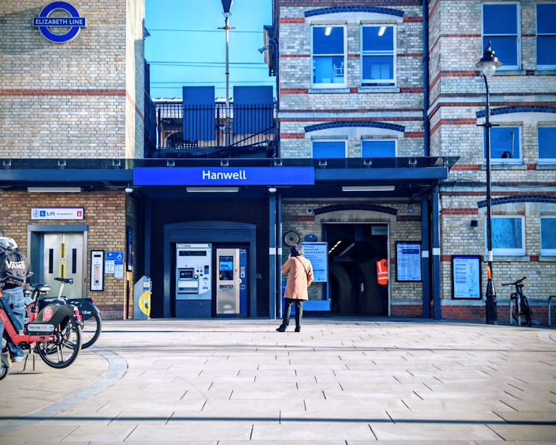 A woman in a beige coat stands outside the entrance to Hanwell station, which features brick construction with a blue sign displaying the station name. In front of the station, on the pavement, there are several bicycles parked, including one with a luggage rack. To the left, a bike-sharing station with branded bikes is visible. The station entrance includes two ticket machines, and to its right, a woman in an orange safety vest appears to be working or monitoring the area near an emergency intercom or information display. The surrounding environment has a clear, sunny atmosphere with a lamppost on the right and building windows above the station entrance, indicating a typical urban setting suitable for home relocation or furniture transport services. Man with Van Shadwell can assist with removals and moving logistics in such areas, handling packing, loading, and transportation efficiently.