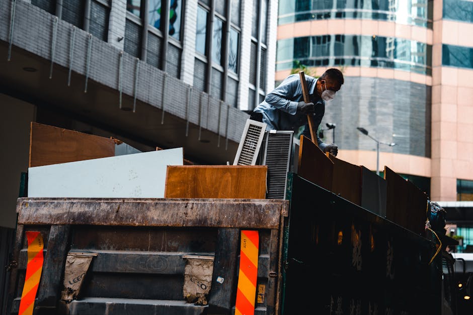 A man wearing a face mask, grey long-sleeved shirt, and gloves is standing on the back of a large black flatbed truck, which is parked on a city street near modern office buildings with glass and concrete facades. The truck is loaded with various discarded household furniture and bulky waste items, including wooden panels, a white appliance or panel, and other miscellaneous materials. The man is lifting or adjusting a piece of furniture, using a wooden stick or crowbar, as part of a home relocation or furniture transport process. The environment is well-lit with natural daylight, and the loading area is adjacent to a building entrance or pavement, indicating an ongoing collection or clearance activity conducted by professional removals or waste disposal services, such as Man with Van Shadwell. The image captures the logistics involved in clearing bulky waste prior to a house move or renovation, highlighting the use of equipment like gloves and strategic positioning of items for efficient loading.