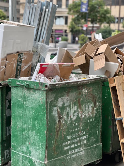 A large, green, and weathered commercial waste dumpster positioned outdoors on a paved area, filled with various discarded items such as flattened cardboard boxes, small packaging materials, and plastic bags. Surrounding the dumpster are partially visible construction debris, including wooden pallets and cardboard packaging. In the background, there are urban elements like a street with parked cars, trees, and buildings, indicating an area of active city environment. The scene depicts the collection of bulky waste, relevant to move-related disposal services in Shadwell, as part of residential or commercial home relocation or furniture transport processes handled by Man with Van Shadwell. The natural lighting suggests daytime with overcast weather, emphasizing the practical aspect of waste management during home moving or removal projects.