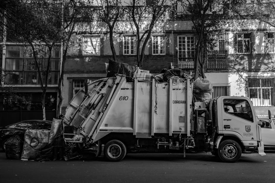 A man wearing a face mask, grey long-sleeved shirt, and gloves is standing on the back of a large black flatbed truck, which is parked on a city street near modern office buildings with glass and concrete facades. The truck is loaded with various discarded household furniture and bulky waste items, including wooden panels, a white appliance or panel, and other miscellaneous materials. The man is lifting or adjusting a piece of furniture, using a wooden stick or crowbar, as part of a home relocation or furniture transport process. The environment is well-lit with natural daylight, and the loading area is adjacent to a building entrance or pavement, indicating an ongoing collection or clearance activity conducted by professional removals or waste disposal services, such as Man with Van Shadwell. The image captures the logistics involved in clearing bulky waste prior to a house move or renovation, highlighting the use of equipment like gloves and strategic positioning of items for efficient loading.