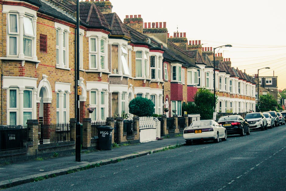 A row of terraced houses in Shadwell, Tower Hamlets, with brick facades and bay windows, some with open window panels revealing curtains inside. The street features parked cars along the pavement, including a white car closest to the foreground. In front of the houses, there are small front gardens with greenery, enclosed by low brick walls and iron railings. Street lamps and utility poles line the pavement, and the sky is overcast, providing soft, diffused lighting. The scene depicts an urban residential area suitable for house removals and furniture transport, with loading areas adjacent to the properties. Occasionally, moving equipment such as trolleys or packing materials may be part of the process, with [COMPANY_NAME], Man with Van Shadwell, providing professional removals and packing services for home relocation. This image reflects the typical environment for local house removals, involving loading and unloading activities that essential moving services facilitate within this neighbourhood.