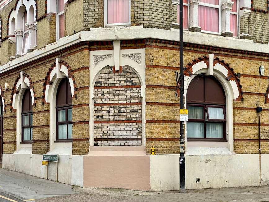 A street view of a multi-storey residential building made of yellow and brown brickwork with arched windows framed in white trim, situated on a corner of a pavement. The building features decorative brick patterns and white architectural details around the windows and on the facade. In front of the building, there is a black lamppost with a small yellow sign attached, as well as a street sign reading 'Chaldon Road'. The sidewalk shows a cardboard box and some packed items suggesting packing for a home relocation. Visible area include a section of a truck or van being loaded with furniture and boxes, and a move blanket or protective padding covering some items being lifted or carried. The scene appears to capture part of a furniture transport or packing process conducted by Man with Van Shadwell, a company that offers removals and house moving services, with a focus on the logistics involved in Shadwell moves, potentially requiring permits for street parking or loading zones.