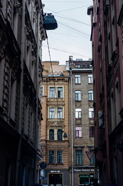 A narrow city street scene showing tall, multi-storey buildings on either side, with a cable car suspended overhead crossing above the street. The buildings are constructed in a historic architectural style with detailed facades, large windows, and decorative elements, and feature a mix of light and dark brickwork. The ground level includes storefronts with signage, referencing local businesses. The street and building facades suggest an urban environment typical for house removals or furniture transport services, with a focus on tight access. Visibility of the street indicates potential logistical considerations for home relocation or moving operations, which [COMPANY_NAME] such as Man with Van Shadwell specializes in managing. The lighting is natural, likely diffused daylight, highlighting the textures of the brickwork and the street scene, providing a clear view of the urban environment suitable for describing the challenges of narrow-access moves.
