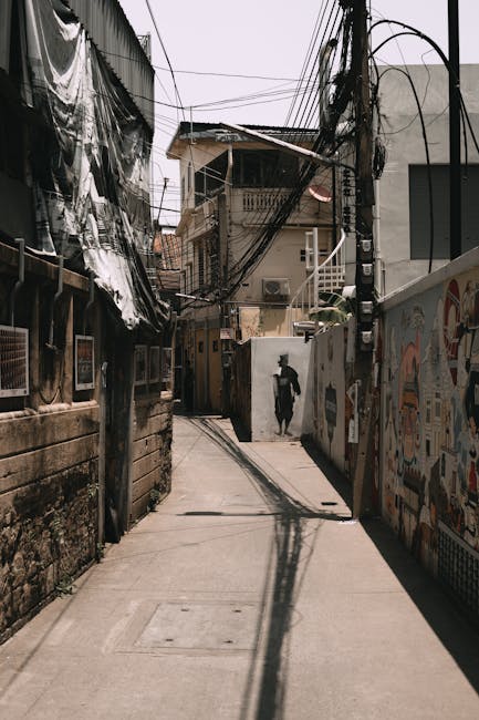 A narrow alleyway in Shadwell with closely positioned residential buildings featuring tiled and concrete facades. A man with van Shadwell is involved in a home relocation, visible loading or unloading a black van parked at the end of the alley, which is partially obscured by the surrounding structures. The alley contains various objects related to moving, including several cardboard boxes, some wrapped in plastic and fabric, placed on the ground and near the entrance of the buildings. A motorcycle is parked along the side of the alley, with a large black bag or wrapped item secured on its seat. Metal scaffolding and electrical wires span across the alley from building to building, with some greenery visible along the edges. The scene is illuminated by natural daylight, capturing the process of furniture transport and packing during a compact urban move, supported by the meticulous handling of boxes and equipment typical in internal property transfers. This image represents the logistical environment involved in residential moves within densely built urban areas like Shadwell.