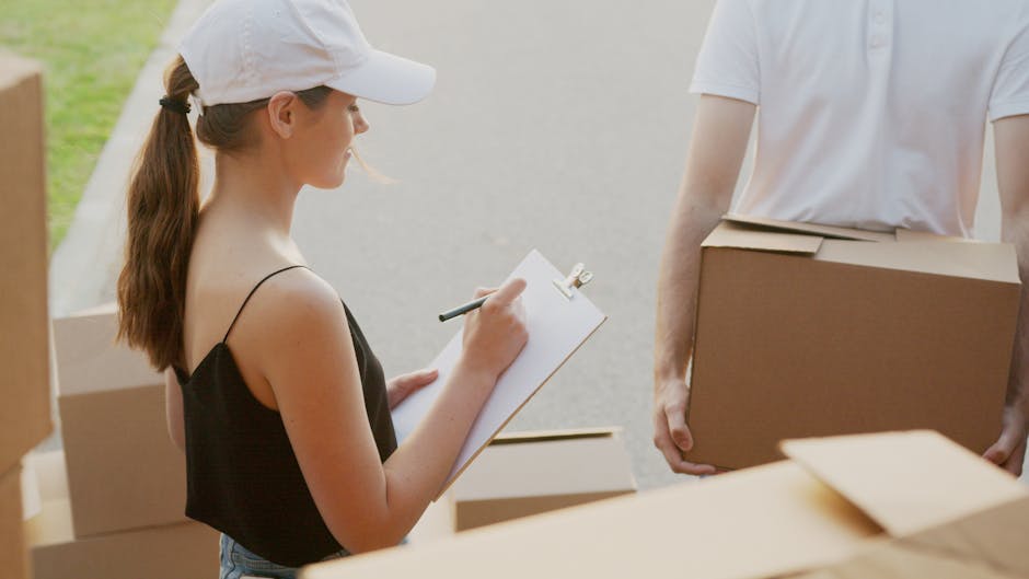 A man wearing a black cap, white t-shirt, and jeans is standing outdoors against a partly cloudy sky, carrying a large cardboard box in both hands. In the background, a vehicle with an open loading ar