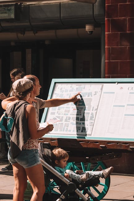 A woman and a young girl are standing outdoors in front of an interactive map display mounted on a wooden stand, located on a pavement near a building with dark brick and metal piping. The woman, wear