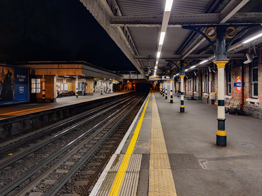 The image depicts the entrance and platform area of Hammersmith underground station, part of the London Underground network, identifiable by the orange and blue roundel logo on the wall. The platform is empty except for a single passenger walking along the tiled floor, which is adjacent to the train tracks. Overhead, there is a large, curved, transparent canopy providing shelter, supported by metal framing. A yellow directional sign with black text indicates the way out toward Talgath Road and bus connections, hanging from the ceiling on the right side of the image. The station’s walls are clad in light-colored panels, and there are advertisement displays visible. The station environment appears clean and well-maintained, with lighting fixtures illuminating the area evenly. This scene represents a typical setting within an urban railway station, relevant for illustrating transit and transportation aspects in a home relocation or moving context, such as loading furniture or packing materials in proximity to rail access.