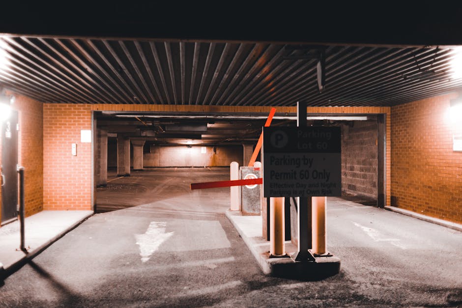 View of a parking garage entrance with a black ceiling and brick walls on either side. In the foreground, there is a parking barrier gate with red and orange arms, a small sign indicating parking lot 60 and permit requirements, and protective bollards. The garage interior is dimly lit, revealing empty parking spaces, concrete support pillars, and a slightly sloped floor leading further into the underground parking area. The environment appears clean and organized, suitable for vehicle access during house removals or furniture transport. The scene reflects a typical setting for loading or unloading items during a home relocation, with an emphasis on parking regulations and safe access for removal vehicles, aligning with the services provided by Man with Van Shadwell.