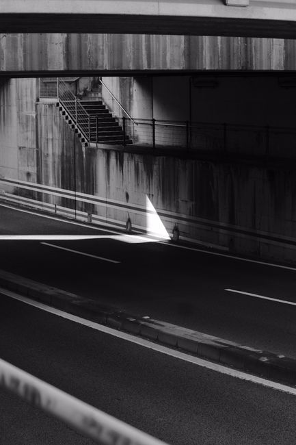 A narrow underpass beneath a concrete building with a staircase on the left side, featuring metal railings and steps, and a roadway with painted lane markings passing through. The scene is illuminated by natural light filtering through an opening, creating a bright triangular patch of light on the road surface. The concrete walls and structure show weathering and stains, and no vehicles or people are visible in the image. This setting could be part of an urban environment where house removals or furniture transport may occur during a home relocation process, with the underpass providing a route for moving vehicles or equipment. The image is in black and white, emphasizing the structural elements and light contrasts.