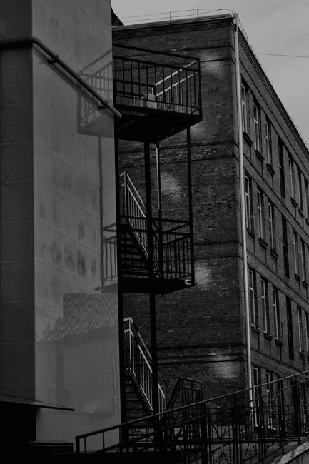 Black and white photograph showing the exterior of a multi-storey brick building on The Highway in Shadwell, with an external metal fire escape staircase attached to the side. The staircase comprises several landings connected by metal steps and railings, with shadows cast on the building's wall. In the foreground, part of a paved street or pavement is visible. The image captures the urban environment and architectural details relevant to house removals and furniture transport, as handled by Man with Van Shadwell, during moving or relocation procedures involving staircases and narrow access points.