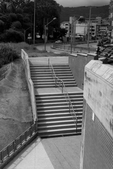 A narrow underpass beneath a concrete building with a staircase on the left side, featuring metal railings and steps, and a roadway with painted lane markings passing through. The scene is illuminated by natural light filtering through an opening, creating a bright triangular patch of light on the road surface. The concrete walls and structure show weathering and stains, and no vehicles or people are visible in the image. This setting could be part of an urban environment where house removals or furniture transport may occur during a home relocation process, with the underpass providing a route for moving vehicles or equipment. The image is in black and white, emphasizing the structural elements and light contrasts.