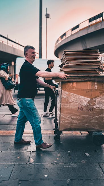 A man in casual clothing, including a black top and blue jeans, is pushing a large trolley loaded with multiple flattened cardboard boxes stacked and wrapped in plastic film. The trolley is positioned on a wet pavement beneath an overpass, with other pedestrians visible in the background. The scene appears to be part of a home relocation or furniture transport process managed by Man with Van Shadwell, with the loading activity taking place outdoors during daylight, close to a bridge structure, illustrating the logistics involved in house removals and packing for moving services.